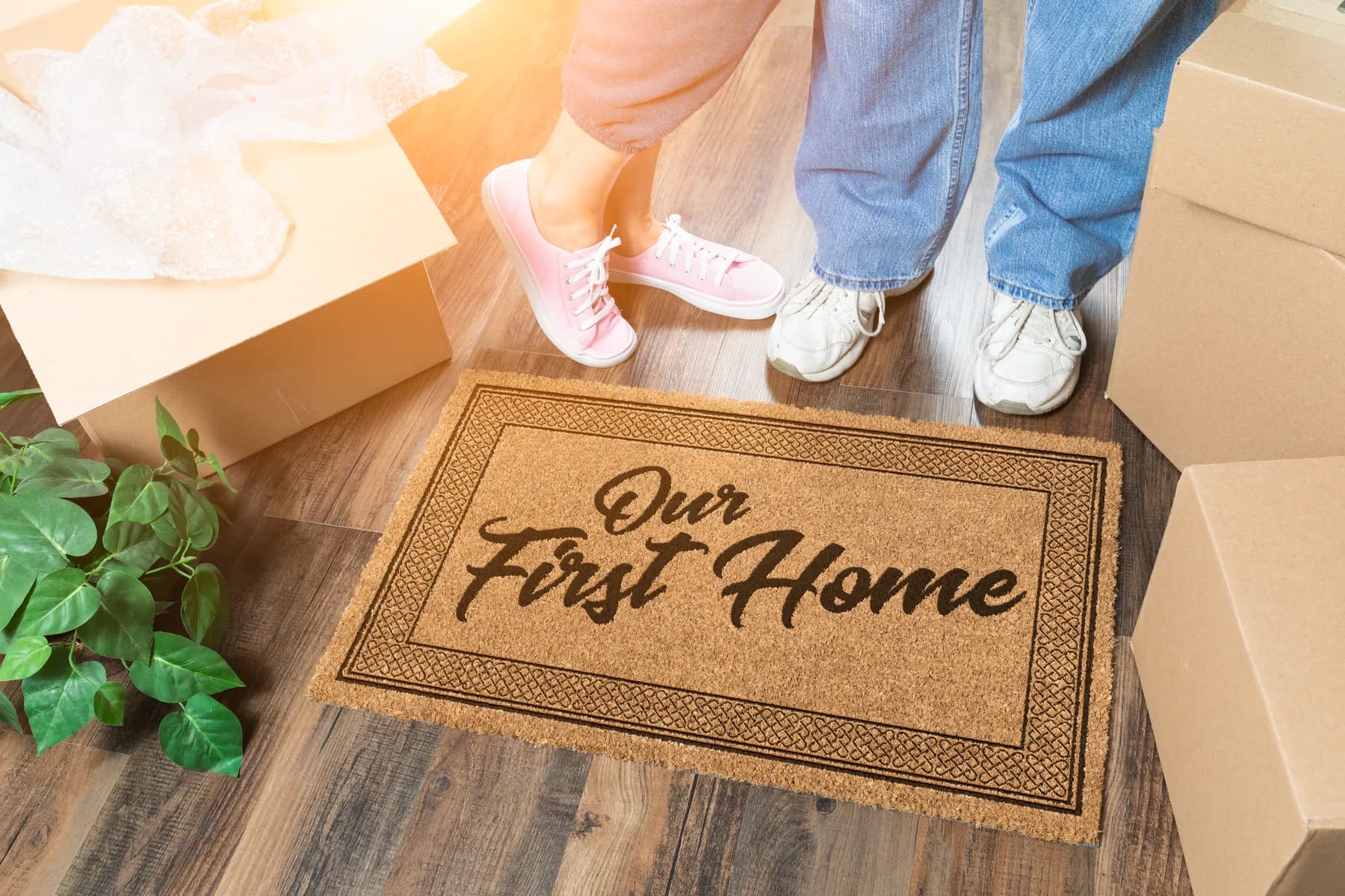 Una pareja parada sobre un tapete que dice “Our First Home”, entre cajas de mudanza